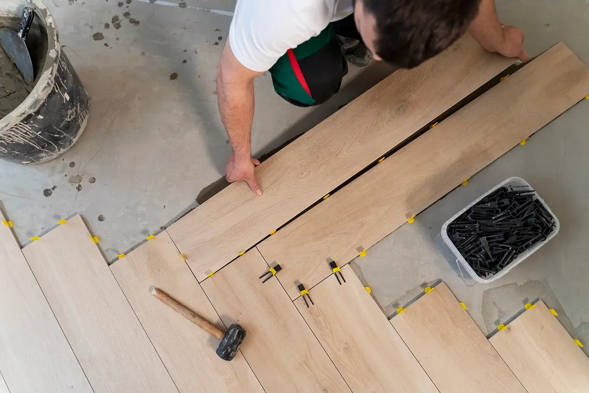 Worker placing wood-style tiles with yellow leveling spacers and a rubber mallet on a concrete floor.