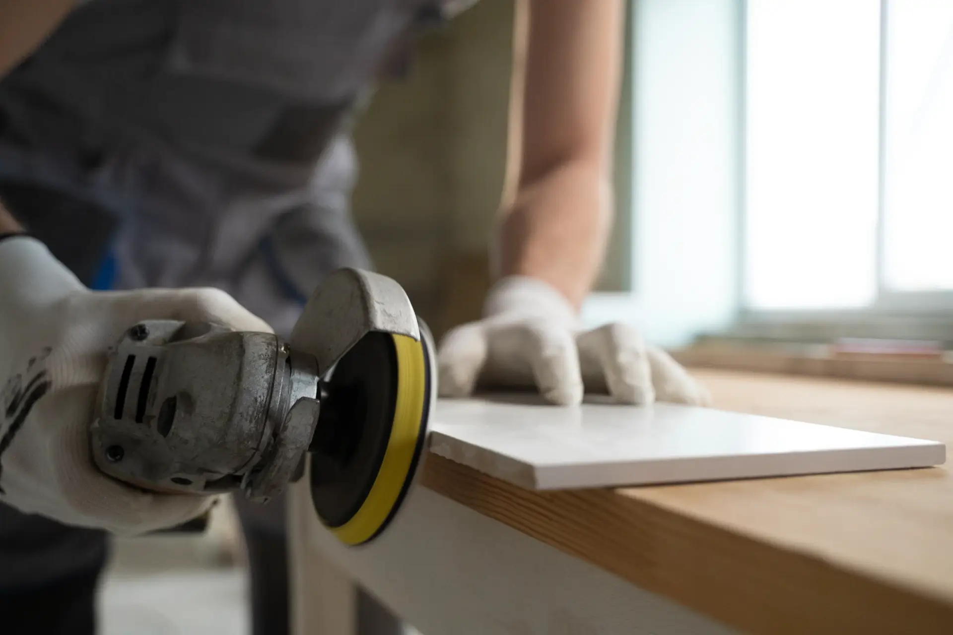 Close-up of a worker in gloves using an angle grinder with a sanding disc to smooth the edge of a white ceramic tile.