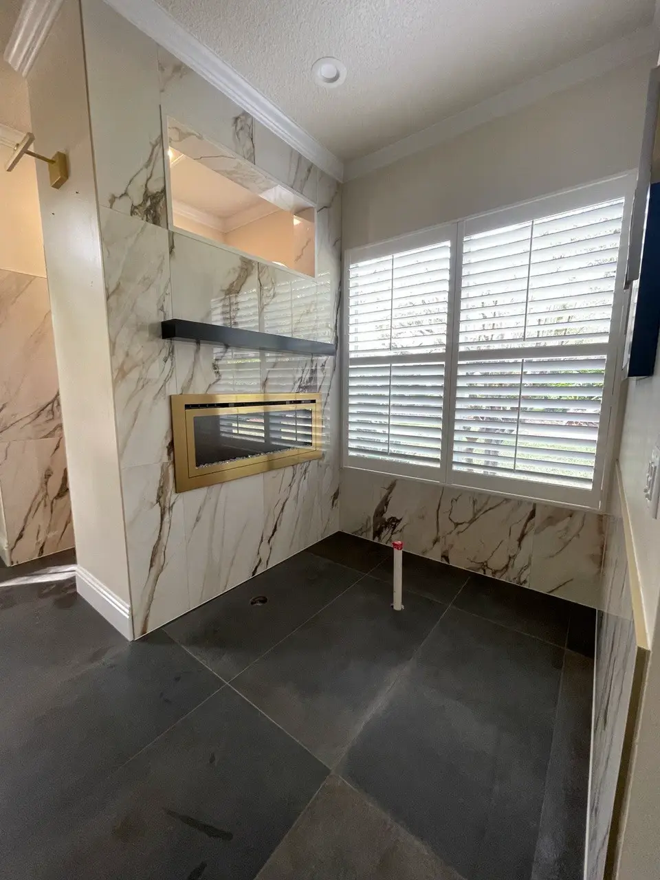 Unfinished modern bathroom with dark floor tiles, white plantation shutters, and a marble-look fireplace surround.