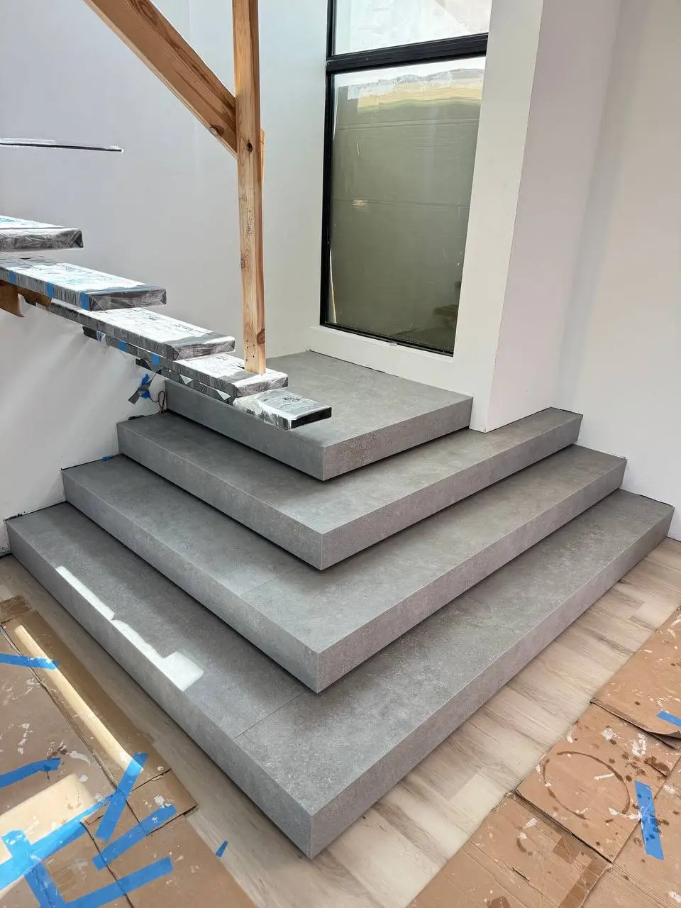 Unfinished modern staircase with a tiered grey stone base and floating steps wrapped in plastic near a tall window.