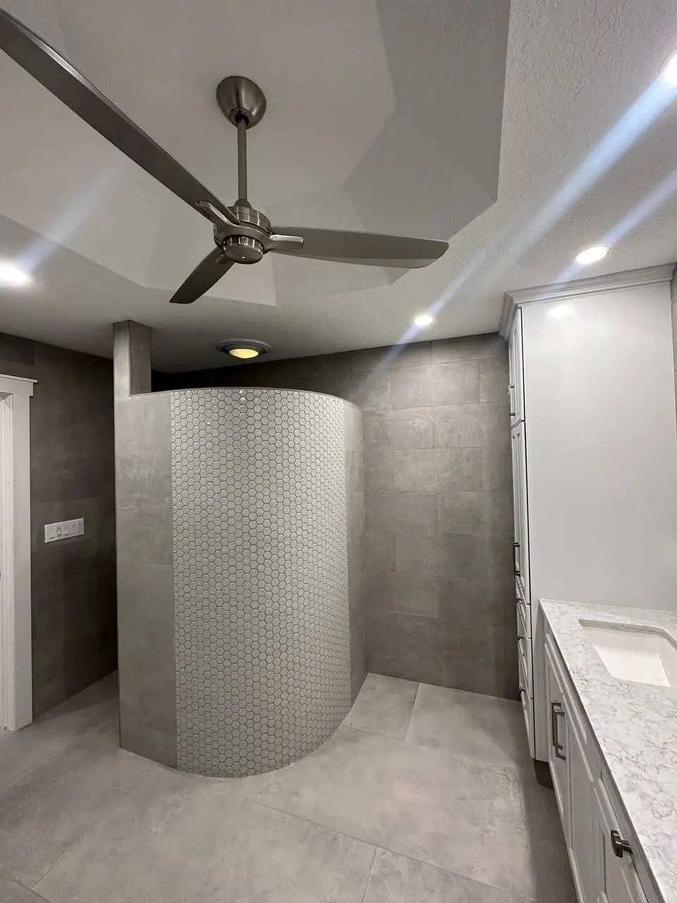 Spacious grey bathroom featuring a curved shower wall with hexagon tiles, a white vanity, and a ceiling fan.