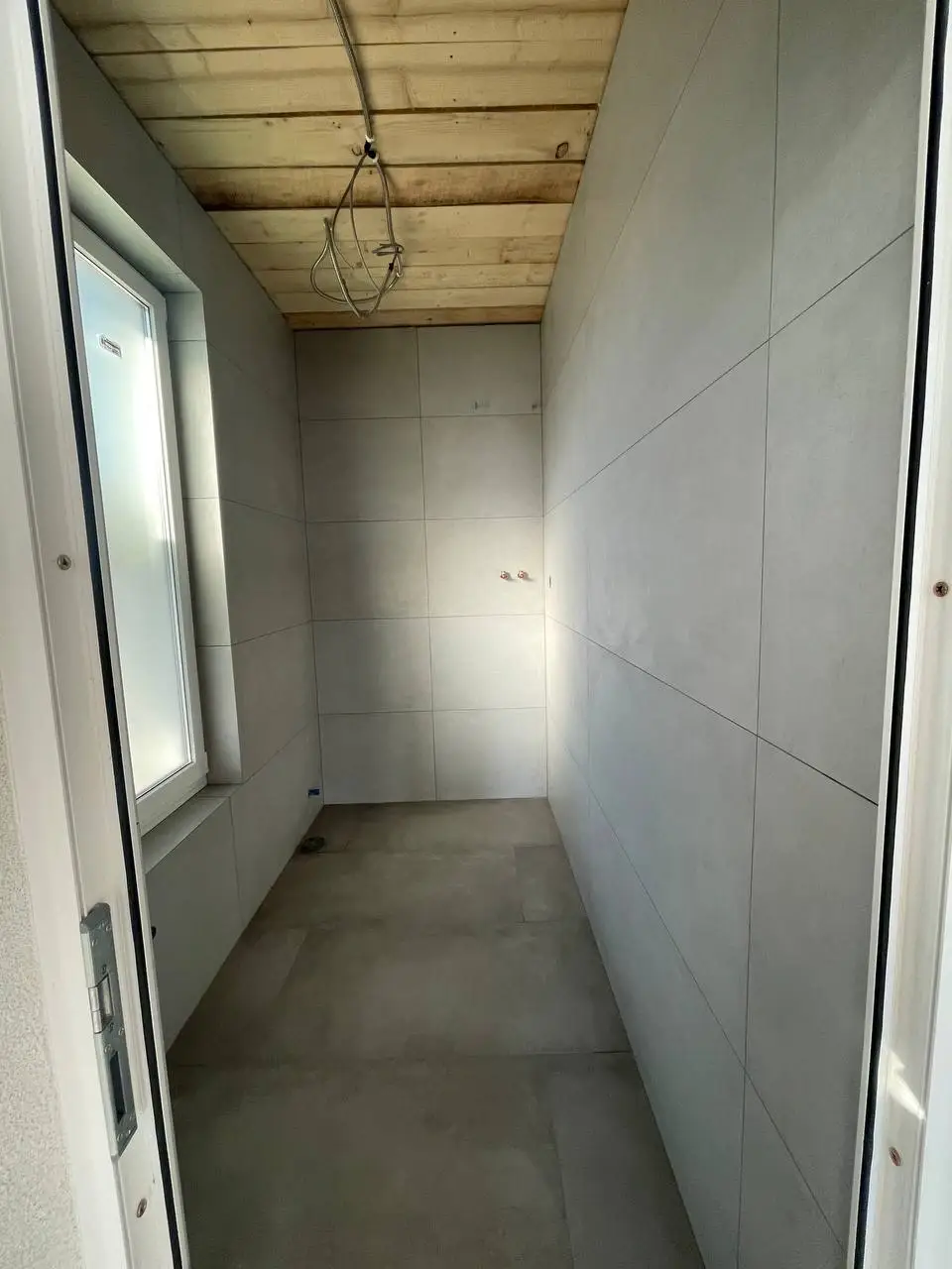 Narrow unfinished bathroom clad in large matte grey tiles with an exposed wood ceiling and hanging wires.