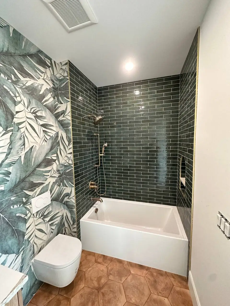 White bathtub surrounded by glossy dark green subway tiles, a palm leaf wallpaper wall, and terracotta hexagon floor tiles.